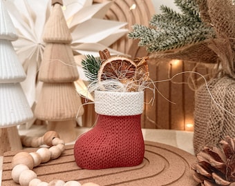 May include: A small red stocking ornament with a white textured cuff, filled with dried orange slices, cinnamon sticks, and a small green pine sprig. The stocking features a detailed knit pattern and is displayed as a decorative holiday item.