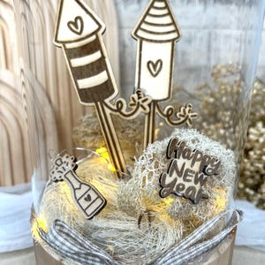 May include: A decorative glass jar with wooden New Year's Eve themed ornaments. The jar contains two rocket-shaped ornaments, a champagne bottle, and a 'Happy New Year' sign. The jar is decorated with a checkered ribbon and fairy lights.