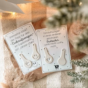 May include: Two white shopping cart tokens with various designs, including a shopping cart, a smiley face, and a heart. Each token is on a white card with German text. The cards are on a wooden surface.