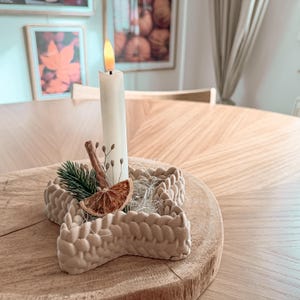 May include: A star-shaped beige candle holder with a textured knit-like pattern. It holds a single white taper candle with a lit flame. The base is decorated with a dried orange slice, a cinnamon stick, a small pine sprig, and dried floral stems.