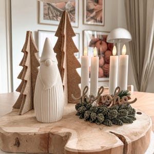 May include: A white gnome figurine with a ribbed base, two tan felt tree cutouts, and four white pillar candles arranged in a circle. A green pine cone wreath base features a brown script sign reading hohoho with dried orange slices.
