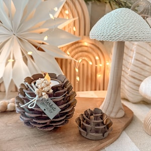 Puede incluir: Decoración en forma de hongo de madera con una parte superior tejida y un tallo de madera, junto a un portavelas en forma de piña. El portavelas tiene una etiqueta que dice "Herbst Geflüster". La decoración está sobre una superficie de madera.