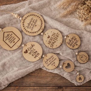 Puede incluir: Adornos decorativos de madera con varias frases y símbolos grabados. Los adornos son redondos y están unidos con hilo y cuentas de madera. Las frases incluyen "Bienvenido" y "El hogar es donde está el corazón."