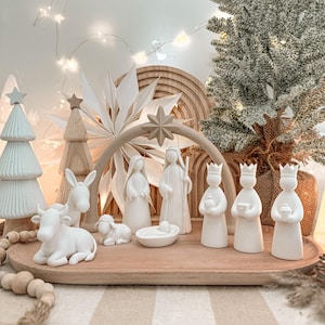 Könnte beinhalten: Ein weißes Krippenset aus Keramik mit stilisierten Figuren von Maria, Josef, dem Jesuskind in der Krippe, drei Königen, einem Esel und einem Schaf. Die Figuren sind auf einem ovalen Holztablett unter einem beigen Bogen mit Stern angeordnet.