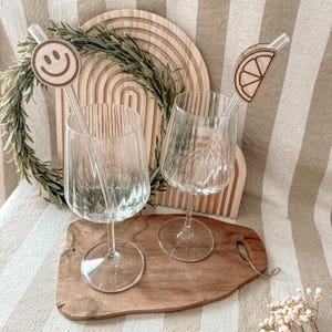 Puede incluir: Dos copas de vino transparentes con detalles acanalados, cada una con un agitador decorativo. Un agitador presenta una carita sonriente de madera, el otro una rodaja de limón de madera. Las copas están sobre una tabla de cortar de madera.