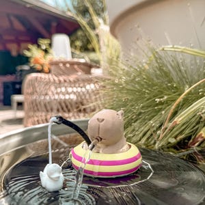 May include: A brown capybara figurine floating in a yellow and pink striped inner tube. The capybara holds a black fishing rod with a white string attached to a small white duck figurine, all positioned on a circular, clear, water-filled surface.