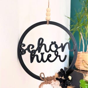 May include: A black circular wall hanging with the German words "schön hier" (beautiful here). The sign is decorated with a small bundle of dried flowers and grasses in neutral tones, suspended by a natural twine.