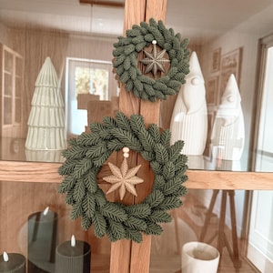 Pode incluir: Duas coroas circulares feitas de agulhas de pinheiro artificiais verde-escuras, cada uma com um ornamento de estrela em tom dourado no centro, suspenso por uma pequena conta de madeira. As coroas estão fixadas na moldura vertical de madeira.