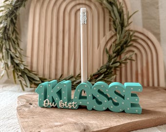 Könnte beinhalten: Ein türkisfarbener Stifthalter mit den Worten "1. Klasse Du bist" in Weiß, der einen Bleistift aus Holz hält. Der Halter hat weiße Tupfen und steht auf einer Holzoberfläche. Ein Holzbogen und Grünzeug sind im Hintergrund.
