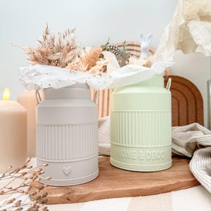 May include: Two decorative milk cans, one light gray and one pale green, sit on a wooden board. The gray can has a heart detail, while the green can has the text "HOME & DECO". Both cans hold dried flowers and are accented with white paper.