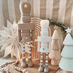 May include: Two decorative nutcracker figurines in beige and white, each with a star-shaped head and star accents. The figurines are adorned with star-shaped details and stand on striped legs. Christmas decor.