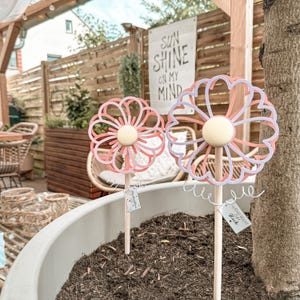 Könnte beinhalten: Zwei dekorative Blumen-Gartenstecker in einem Pflanzgefäß. Die Blumen sind rosa und weiß mit einem gelben Mittelteil, befestigt an Holzstäben. Das Pflanzgefäß ist mit Erde gefüllt. Ein Schild im Hintergrund lautet "SUN SHINE ON MY MIND."