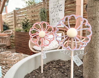 May include: Two decorative flower garden stakes. Each flower has a layered design with pink and white petals and a yellow center. The stakes are in a large gray pot filled with soil. A banner in the background reads "SUN SHINE ON MY MIND."