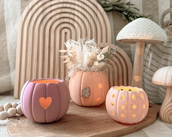 May include: Three pumpkin-shaped candle holders in shades of pink and orange, with heart and dot cutouts. One pumpkin has a leaf accent. The candle holders sit on a wooden board with a wooden arch and mushroom decor in the background.