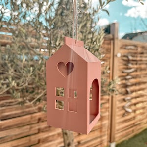 Könnte beinhalten: Ein rosafarbenes Vogelhaus in Hausform, das an einer Schnur hängt. Es hat einen herzförmigen Ausschnitt und quadratische Fensterausschnitte. Der Hintergrund zeigt einen Baum, einen Holzzaun und einen teilweise bewölkten Himmel.