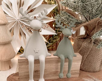 Puede incluir: Dos figuras de renos con patas largas y astas sentadas sobre un bloque de madera rectangular. El reno más grande es de color gris claro con una pequeña estrella en el pecho, mientras que el más pequeño es de color verde salvia. Ambos llevan un cordel.