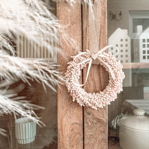 Op de afbeelding: Een kleine, ronde krans bestaande uit talloze piepkleine, lichtroze besachtige bolletjes. De krans hangt aan een lichtbeige stoffen lint en is voorzien van een klein, centraal houten hartje dat is vastgemaakt met een fijn wit strikje.