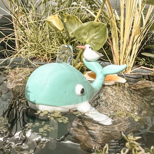 Puede incluir: Una fuente con forma de ballena verde menta claro con vientre blanco y un pequeño pájaro blanco de pico naranja posado en su cola. La ballena tiene un ojo grande blanco con pupila negra y agua brota de la parte superior de su cabeza al estanque.
