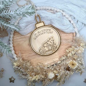 Puede incluir: Adorno navideño de madera con el texto "Oh du schöne Weihnachtszeit" y un camión que transporta un árbol de Navidad. El adorno forma parte de una corona con flores secas, vegetación y decoraciones en forma de estrella.