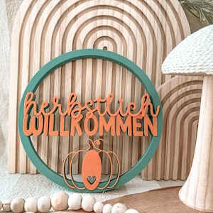 May include: A teal wooden sign with the German words "herbstlich WILLKOMMEN" in orange script, and an orange pumpkin graphic with a heart cutout. The sign is set against a wooden arched background and a mushroom-shaped decoration.