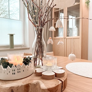 Puede incluir: Una decoración de mesa con un jarrón de cristal transparente con ramas y adornos colgantes de flores blancas. Un portavelas de cerámica blanca con diseño de pueblo y velas encendidas sobre una mesa de madera. También hay portavelas de cristal transparente y posavasos de madera.