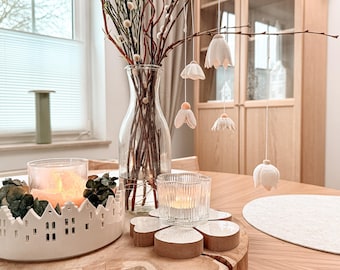 May include: A decorative table setting featuring a clear glass vase with branches and hanging white flower ornaments. A white ceramic candle holder with a village design and lit candles are on a wooden table. A clear glass candle holder and wooden coasters are also present.
