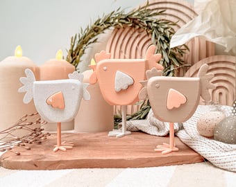May include: Three wooden chicken figurines in white, peach, and tan, each with a contrasting wing and belly. They stand on a wooden board, with candles and decorative wooden circles in the background. The scene evokes a rustic, springtime theme.