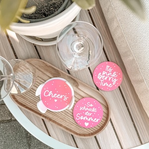 May include: A wooden tray holds two wine glasses and three pink coasters. The coasters have white text that reads "Cheers", "It's pink berry time", and "So schmeckt der Sommer". The coasters are round with white polka dots.