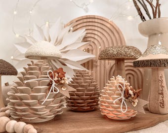May include: Three pinecone-shaped decorative items with light-coloured scales, each adorned with a small white string bow and dried flowers. One pinecone glows from within. Nearby are wooden mushroom figures, one with the text "Glückspilz".