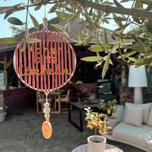 May include: A circular hanging decoration featuring vertical pink stripes and the text "Ab hier bitte lächeln" in gold lettering. The piece is suspended by a string with wooden beads and a small orange smiley face charm at the bottom.