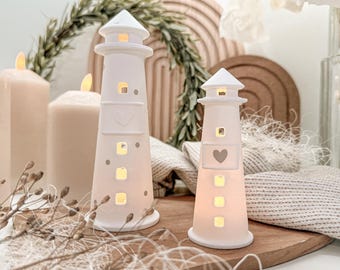 May include: Two white ceramic lighthouse decorations with heart cutouts and square windows. The lighthouses are illuminated from within, creating a warm glow. The lighthouses are on a wooden surface, with candles and decorative elements in the background.