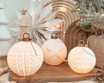 May include: Three spherical white ornaments with a textured knit pattern. Each ornament features a brown loop at the top with a small gold bell attached. The ornaments are illuminated from within, casting a warm glow through the white material.