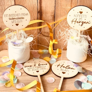 Op de afbeelding: Vier ronde houten borden met verschillende spreuken erop. De borden staan op stokjes en zijn omringd door confetti. De spreuken zijn "Ein bisschen mehr täterätáte uns guti", "Konfetti im Herzen", "in saus & braus wirds närrisch in jedem haus", and "Helau again".