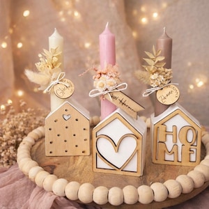 May include: A decorative arrangement featuring three candles in cream, pink, and brown, adorned with dried floral accents and wooden tags. The candles are set on wooden house-shaped holders with heart and HOME cutouts, displayed on a wooden tray with a beaded edge.