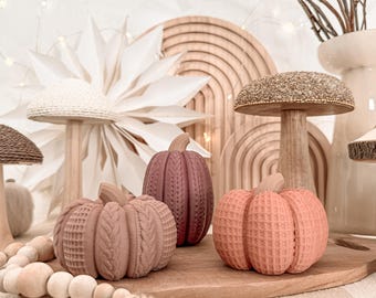 May include: Three decorative pumpkins in shades of mauve, plum, and peach, each featuring a distinct textured surface resembling knit fabric and waffle patterns. The pumpkins are arranged alongside wooden mushroom figures and a string of wooden beads.