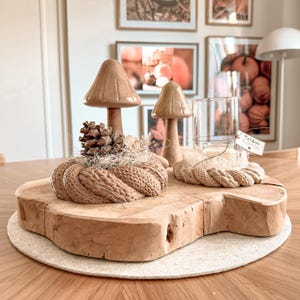 Peut inclure: Un centre de table décoratif composé de deux champignons en céramique, d'une pomme de pin et d'un bougeoir en verre sur une base en tranche de bois naturel. Une corde tressée beige entoure la base des champignons et du bougeoir, avec une petite étiquette portant l'inscription "Herbst KLOPPEN".