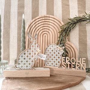 Può includere: Decorazioni pasquali in legno con un coniglio di vimini bianco, un uovo e un cartello che dice "Frohe Ostern".