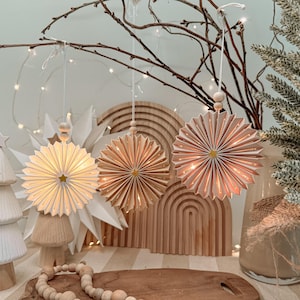 May include: Three circular hanging ornaments made of pleated paper in shades of white, light tan, and soft pink. Each ornament features a small gold star at the center, a wooden bead on the hanging string, and internal lights that create a warm glow.