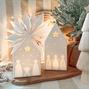 Op de afbeelding: Twee witte keramische kerstversieringen in de vorm van een huis met steruitsparingen en silhouetten van figuren. De versieringen staan op een houten plank, met een papieren ster en een kleine kerstboom op de achtergrond.