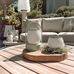 Op de afbeelding: Twee witte, geribbelde lantaarns met zwarte rand staan op een houten oppervlak. Eén lantaarn heeft een groene handgreep en basis met een hartdetail. De andere is omgeven door groen. De scène speelt zich buiten af.