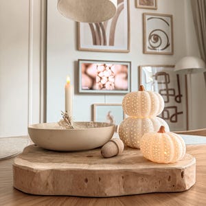 May include: A light-colored wooden slab serving as a base for a beige bowl containing a lit taper candle and dried botanicals, alongside a small wooden acorn and two stacked, illuminated, white ceramic pumpkins with a perforated dot pattern.