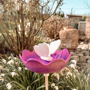 Op de afbeelding: Een tuinpaal met een witte en paarse vlinder. Het heeft een paarse bloemvormige schaal met een witte vlinder erop. De vlinder heeft witte vleugels en een paars lichaam. De paal staat in een tuin met witte bloemen en groen.