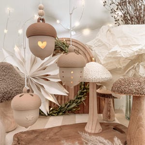 Puede incluir: Tres adornos colgantes con forma de bellota en tonos beige y marrón mate, con recortes en forma de corazón, estrella y pequeños puntos. Cada adorno cuelga de un cordón blanco con cuentas de madera. Debajo hay dos figuras con forma de seta.