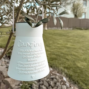 May include: White camping lantern with embossed text including the word "Camping". The lantern hangs from a tree branch with green leaves. The background is a blurred outdoor scene with a house and grass.