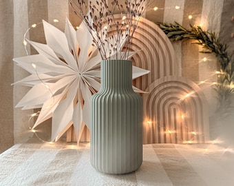 May include: A sage green ribbed vase holds dried floral stems. A large white paper star and wooden arch decorations are in the background. The scene is lit with warm fairy lights, creating a cozy, neutral-toned aesthetic.