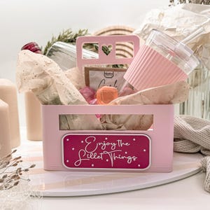 May include: A pink gift basket with a handle and a heart-shaped cutout. The basket contains a clear tumbler with a pink silicone sleeve, a small bottle, and other items wrapped in tissue paper. The basket has a label that reads "Enjoy the Lillet Things."