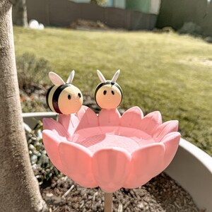 May include: A pink flower-shaped bird bath with two bee figurines. The bees are yellow, black, and white. The bird bath is made of plastic and is placed in a garden setting.