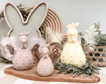 May include: Three decorative ceramic rooster figurines in varying sizes and colors, arranged on a wooden board. The roosters are light pink and yellow, with textured surfaces and feather accents. The background includes a bunny-shaped frame and decorative elements.