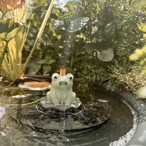 May include: A light green frog figurine with a small orange crown on its head, positioned on a circular black solar panel base. The frog is designed as a water fountain, with a steady stream of water flowing from its mouth into the surrounding pool.