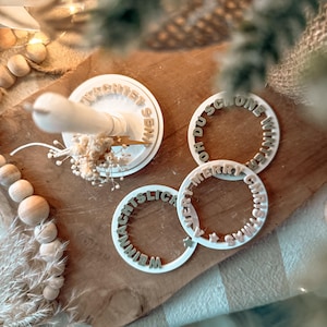 Puede incluir: Cortadores de galletas circulares blancos con texto en relieve, incluyendo "Merry Christmas" y "Oh Du Schone Winter". Los cortadores están dispuestos sobre una superficie de madera, con un arreglo floral decorativo y cuentas de madera.
